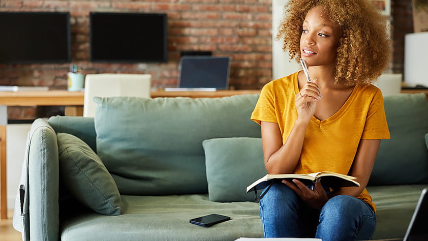 Woman sitting on couch with a pen and notebook, thinking while looking out window