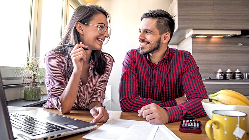 Couple sitting at their kitchen table using a laptop and looking at paperwork