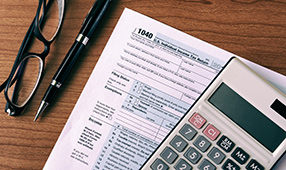 Calculator, a pair of glasses, a pen and tax forms on a wooden table