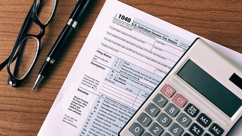 Calculator, a pair of glasses, a pen and tax forms on a wooden table