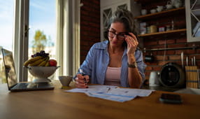 Middle-aged educator adjusts her glasses while reviewing bills, laptop and receipts on the kitchen table, focused on budgeting and when to use a personal loan