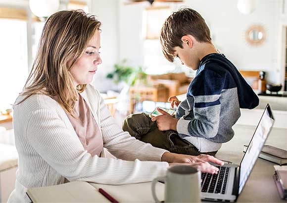 Woman working from home on laptop while son uses smartphone
