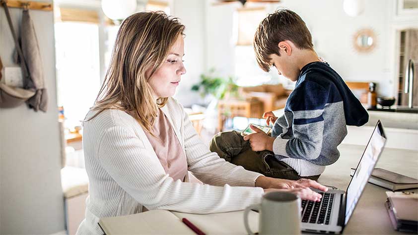 Woman working from home on laptop while son uses smartphone
