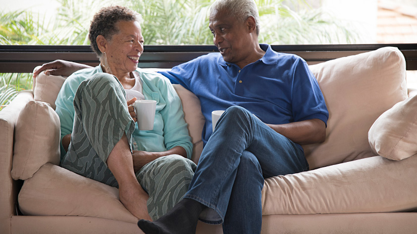 Senior couple sitting on a tan sofa, drinking a hot drink and having a conversation