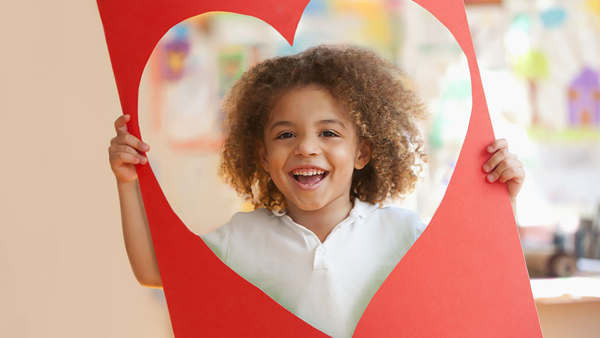 Celebrating Teacher Appreciate Week - Grinning Young Boy Looking Through a Large Red Construction Paper Heart-Shaped Cut-Out