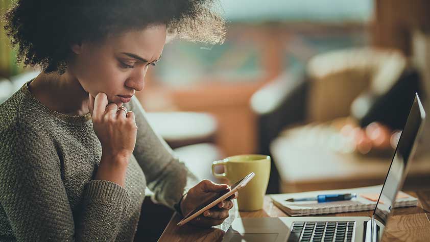 4 Student Loan Scam Red Flags - Concerned African American Woman Using Her Cell Phone While Working on Her Laptop