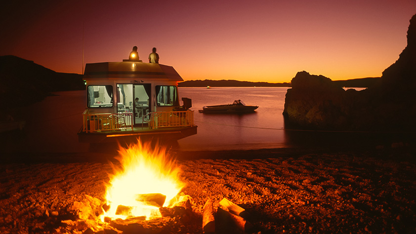 Houseboat camper at sunset with a campfire in foreground and a water-ski boat in the background