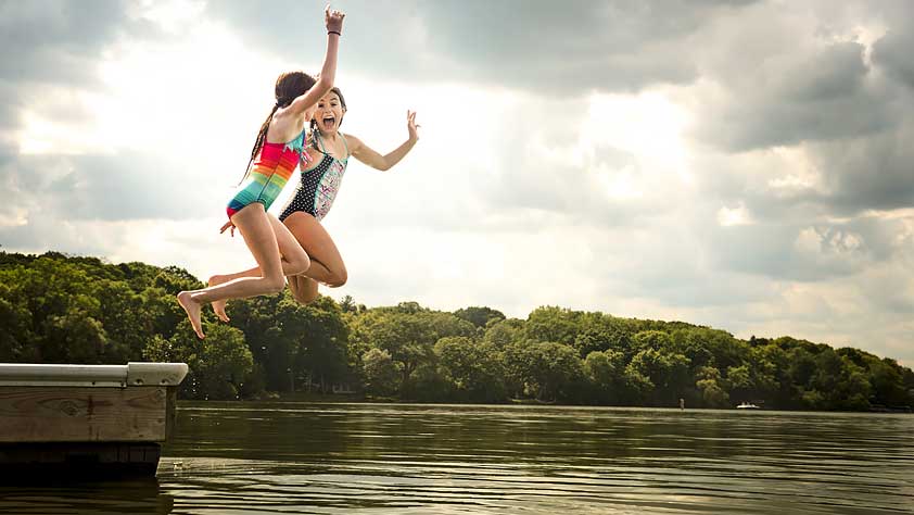 Two Young Girls in Swimsuits Jumping off a Dock into a Lake