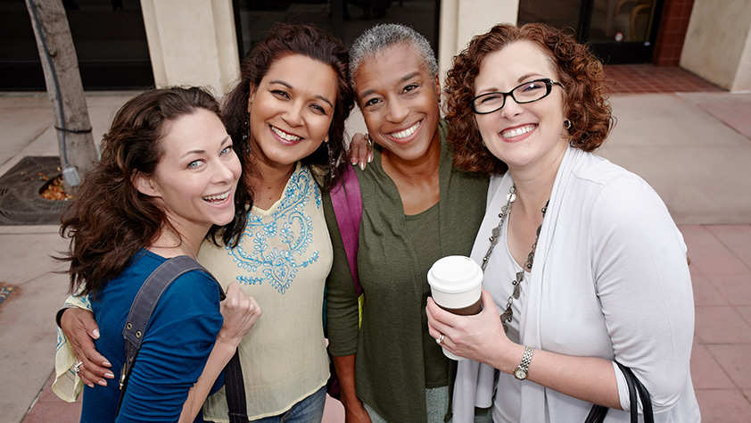 Group of four smiling women standing on a city sidewalk