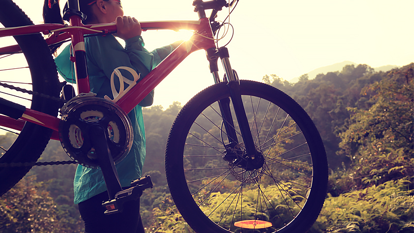 Woman carrying her mountain bike toward a sunrise ride on a wooded trail