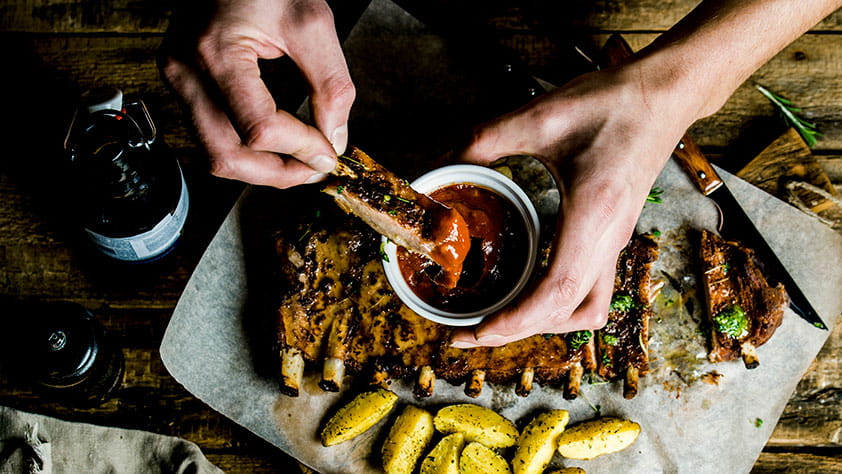 Top view of a man's hands dipping a pork rib into barbecue sauce on rustic wooden table