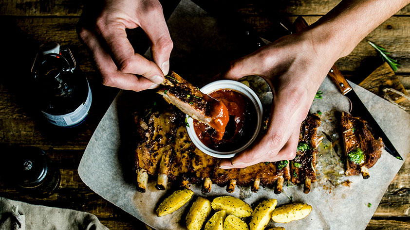 Top view of a man's hands dipping a pork rib into barbecue sauce on rustic wooden table