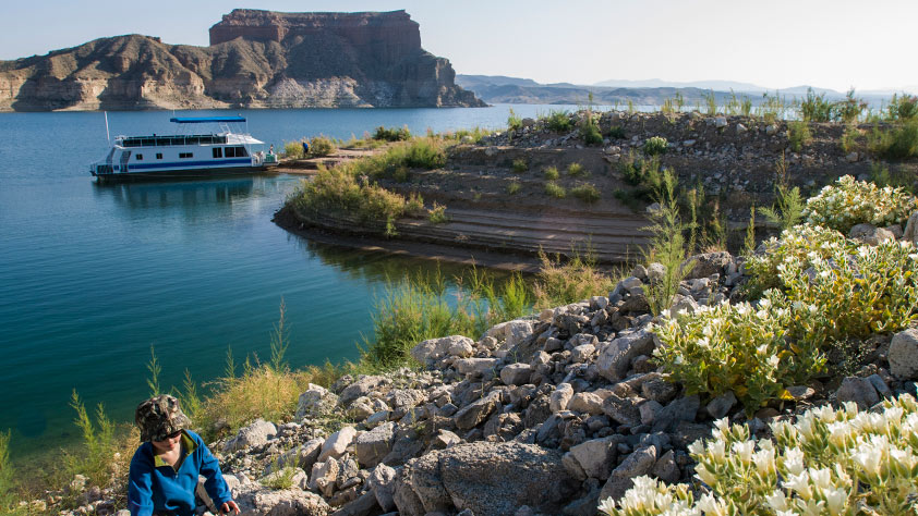 A young boy exploring the shores of Lake Mead with a house boat in the background