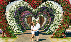Couple holding hands while standing under a heart-shaped sculpture made of live flowers