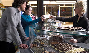 Woman behind a counter in a chocolate shop handing a couple a box of chocolates