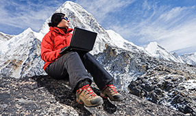 Woman with Laptop Wearing an Orange Parka Sitting on Mountain Rocks