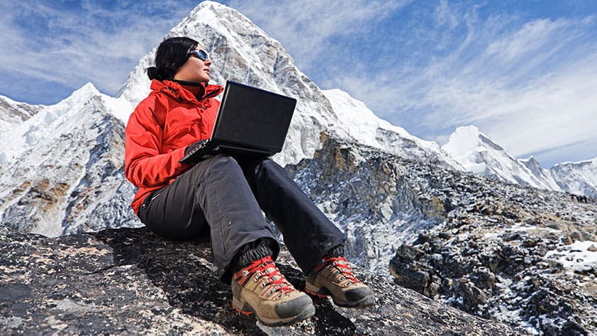 Grants for Educators - Woman with Laptop Wearing an Orange Parka Sitting on Mountain Rocks