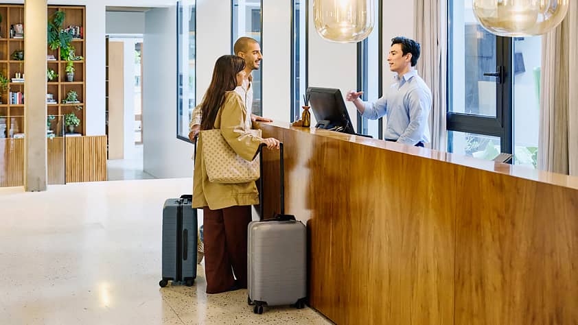 Wide shot of young couple with suitcases at the hotel lobby front desk, checking into a modern hotel.