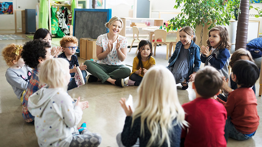 Preschool teacher and students clapping in a circle on the floor in a classroom