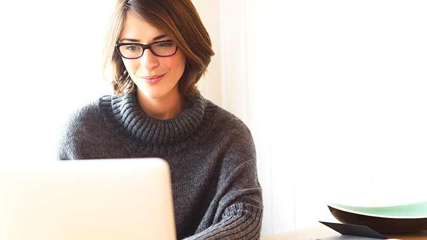 Brunette woman wearing glasses sitting at a table and using her laptop