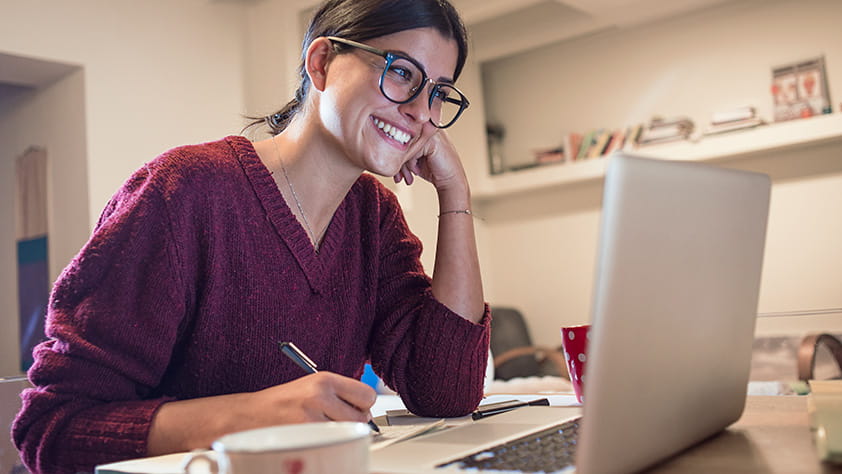 Young woman at home using her laptop and writing in a notebook
