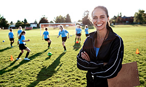 Confident female coach and her middle school girls' soccer team running drills at practice on a sunny field