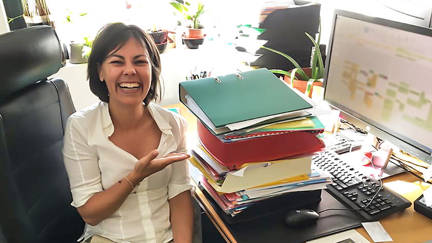 Woman Laughing In Front of Large Stack of Papers on Desk