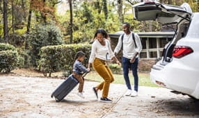 Mother pulling young boy on suitcase and loading car for family vacation