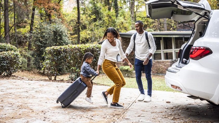 Mother pulling young boy on suitcase and loading car for family vacation