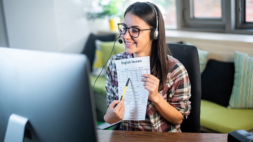 Teacher seated in front of computer at home wearing headset and sharing printed lesson through webcam