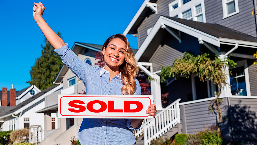 Happy woman with one arm raised and holding a red SOLD sign in the other, standing in front of a row of suburban houses