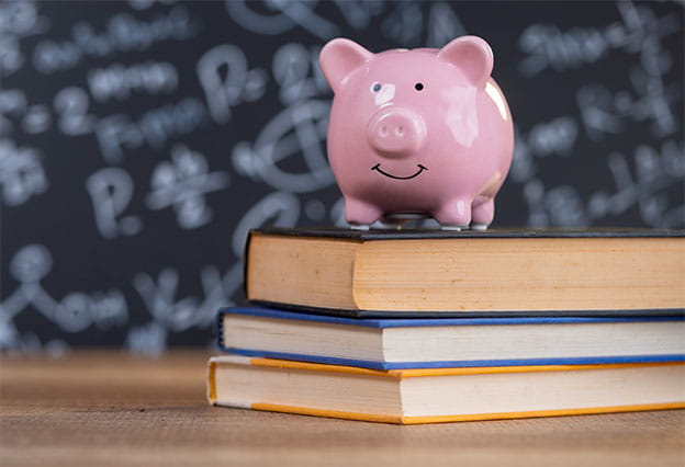 A happy piggy bank sitting on top of a stack of books set upon a school desk.