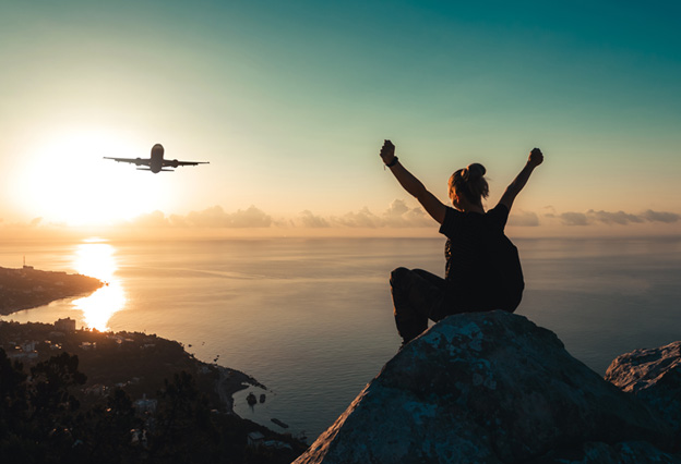 NEA Travel: Airfare - Hiker sitting on the edge of a mountain peak triumphantly reaching both arms up in the air as an airplane flies over the sea coast at sunrise.