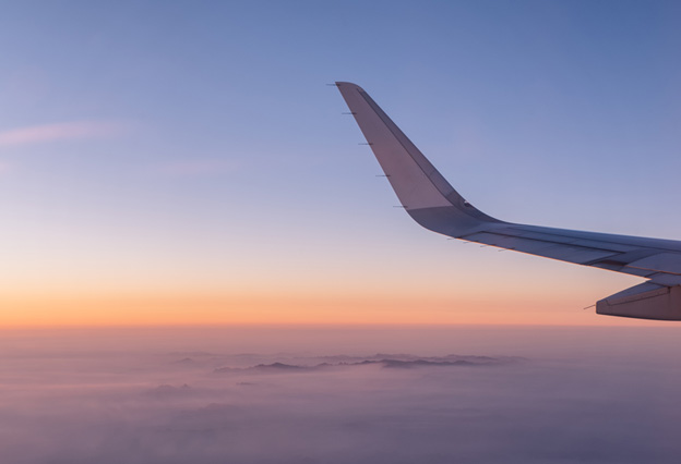 NEA Travel: Airfare - A photo of an airplane wing taken while flying above the clouds during sunset.