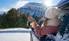 NEA Travel: Car Rental - Young woman renting a car in winter on snowy mountain roads lined with pine trees