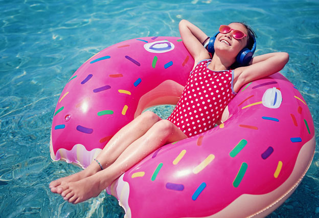 NEA Travel: Resorts - Little girl is enjoying the sunshine on a bright doughnut-themed raft while floating in the water and listening music.