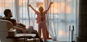 La niña abre las cortinas de una gran ventana panorámica e ilumina la habitación con la cálida luz del atardecer