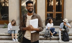 Feliz joven estudiante universitario con un libro parado al aire libre frente al campus